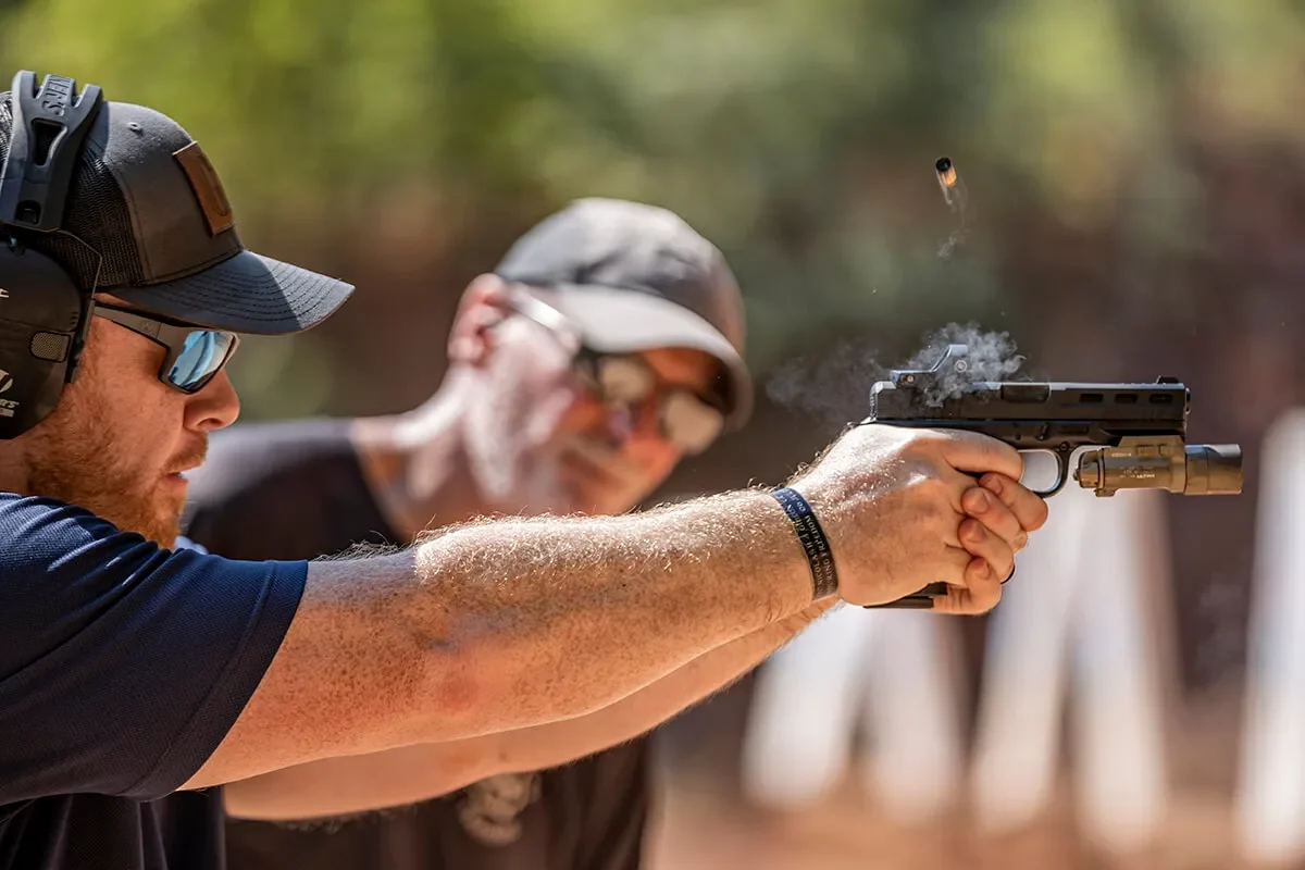 Mężczyzna w słuchawkach ochronnych strzela z pistoletu na strzelnicy – dynamiczne ujęcie z wyrzutem łuski.